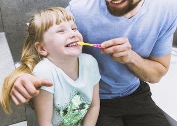 A father is helping his daughter brush her teeth to maintain oral hygiene