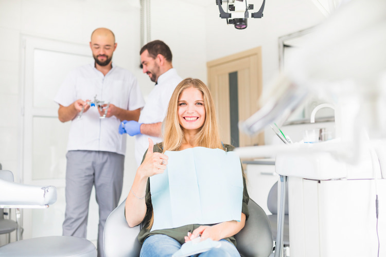 a happy patient sitting on a dental chair while two dentist are talking behind her.